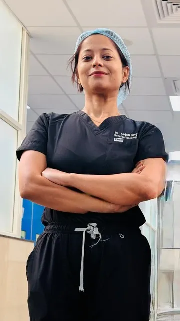 Dr Anjali Singh - Surgical Oncologist Female medical professional in black scrubs with a blue surgical cap, standing in a clinic hallway with arms crossed.