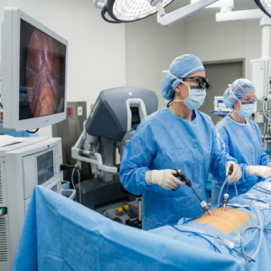 Two surgeons in blue scrubs, masks, and caps perform an operation while viewing a monitor displaying the patient’s internal view.